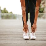 From below crop slender female athlete in sportswear and white sneakers doing standing forward bend exercise for stretching body on wooden floor of street sports ground against blurred urban environment in daytime